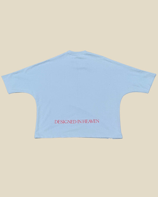 KAIROS EN SU TIEMPO PERFECTO blue tee back with DESIGNED IN HEAVEN embroidery in red text on the lower back, displayed flat against a solid beige background.